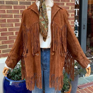 Puede incluir: Una chaqueta de ante marrón con flecos largos, usada sobre una blusa blanca y vaqueros azules. Una bufanda estampada está atada alrededor del cuello. La chaqueta se exhibe en un maniquí frente a una pared de ladrillos.