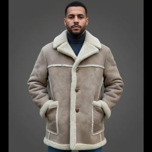 Puede incluir: Un abrigo de piel de oveja de color beige con forro y ribete de piel de oveja color crema. El abrigo tiene una parte delantera abotonada, dos bolsillos delanteros y un cuello ancho. El modelo lleva un cuello alto azul marino.