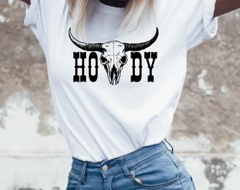 Puede incluir: Camiseta blanca con un gráfico de calavera de toro negro y la palabra "HOWDY". La modelo lleva una gorra de béisbol blanca con una estrella negra y vaqueros azules. Ropa informal de temática occidental.