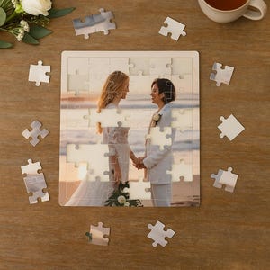 May include: A jigsaw puzzle depicting a wedding photograph of two people holding hands on a beach. Loose puzzle pieces, a cup of tea, and floral decorations surround the puzzle. The image is set on a wooden surface.