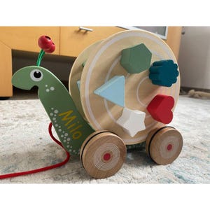 Può includere: Giocattolo in legno da trascinamento a forma di lumaca, con corpo verde e guscio beige. Il guscio presenta ritagli per forme geometriche colorate. Il corpo della lumaca ha il nome "Milo" stampato. Il giocattolo ha ruote in legno e un cordino rosso.