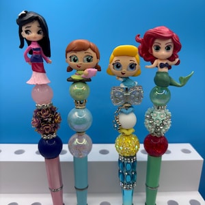 May include: Four decorative pens featuring character toppers. The first has a black-haired figure, pink, purple floral, and blue beads. The second has a brown-haired figure, green, blue, and white beads. The third has a blonde figure, bow, and yellow beads. The fourth has a red-haired mermaid figure, floral, and red beads.