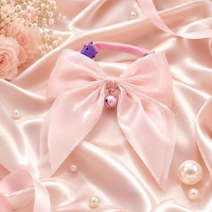 May include: A light pink cat collar with a large, sheer bow and a small bell charm. The collar is set against a backdrop of pink satin fabric, with scattered pearls and floral accents.