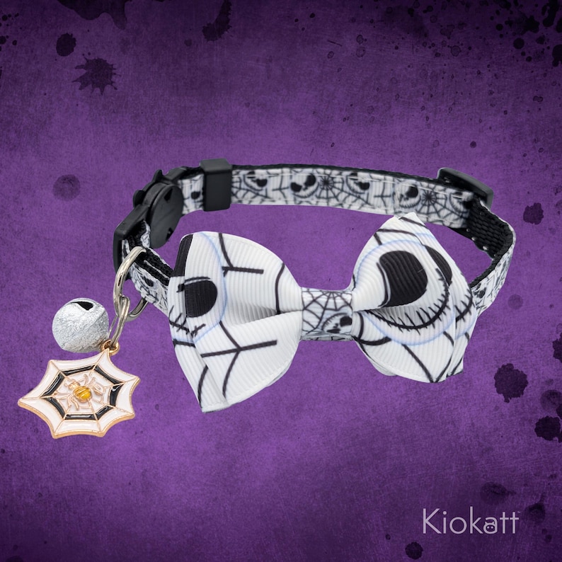 May include: A black and white cat collar with a bow tie featuring a Jack Skellington design. The collar has a matching pattern of spiderwebs and faces. A small bell and spiderweb charm are attached. The brand name "Kiokatt" is at the bottom.