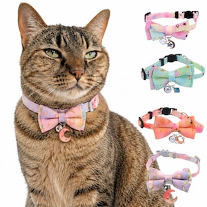 May include: A brown tabby cat wearing a pastel-colored collar with a bow tie and a crescent moon charm. Four additional collars are displayed separately, each featuring a bow tie, a small bell, and a unique celestial-themed metal charm.
