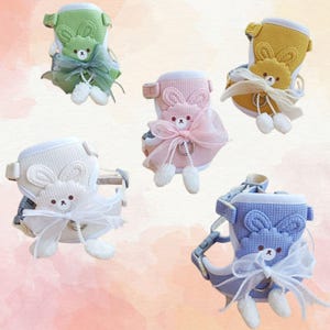 May include: Five pet harnesses in various pastel colors: green, pink, yellow, white, and blue. Each harness features a bunny face design, a bow, and dangling pom-poms. The harnesses are designed for small pets.