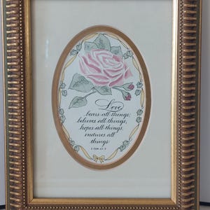May include: A framed print featuring a pink rose illustration and the text: Love bears all things, believes all things, hopes all things, endures all things. 1 Cor 13:7. The artwork is enclosed in an oval mat within a textured gold-toned frame.