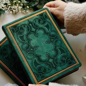 May include: Two green books with gold edging and detailed embossed patterns. The books are stacked, with the top one angled, showing the gold trim. The background features greenery and a soft, neutral surface.