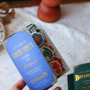 Pode incluir: Uma etiqueta de di&aacute;rio azul com letras douradas que diz "Little Book of Good Vibes Journal Tabs". A etiqueta inclui 60 marcadores de p&aacute;gina autocolantes. Autocolantes decorativos s&atilde;o vis&iacute;veis.