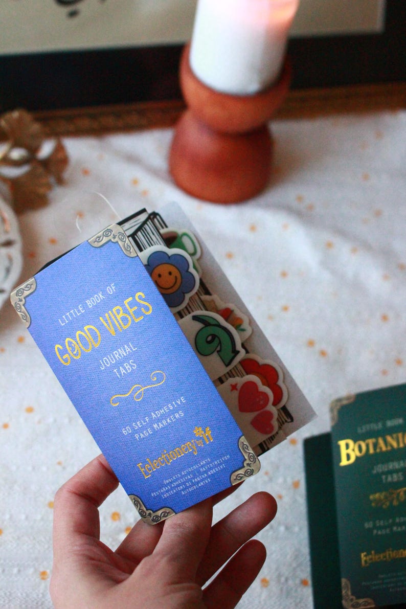 Pode incluir: Uma etiqueta de di&aacute;rio azul com letras douradas que diz "Little Book of Good Vibes Journal Tabs". A etiqueta inclui 60 marcadores de p&aacute;gina autocolantes. Autocolantes decorativos s&atilde;o vis&iacute;veis.