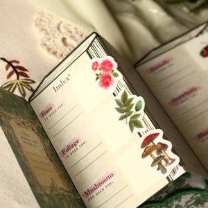 Pode incluir: Di&aacute;rio aberto com adesivos decorativos de flores e cogumelos. A p&aacute;gina do &iacute;ndice apresenta as palavras "Rose", "Foliage" e "Mushrooms" com linhas para anota&ccedil;&otilde;es. O di&aacute;rio est&aacute; aberto em uma superf&iacute;cie creme.