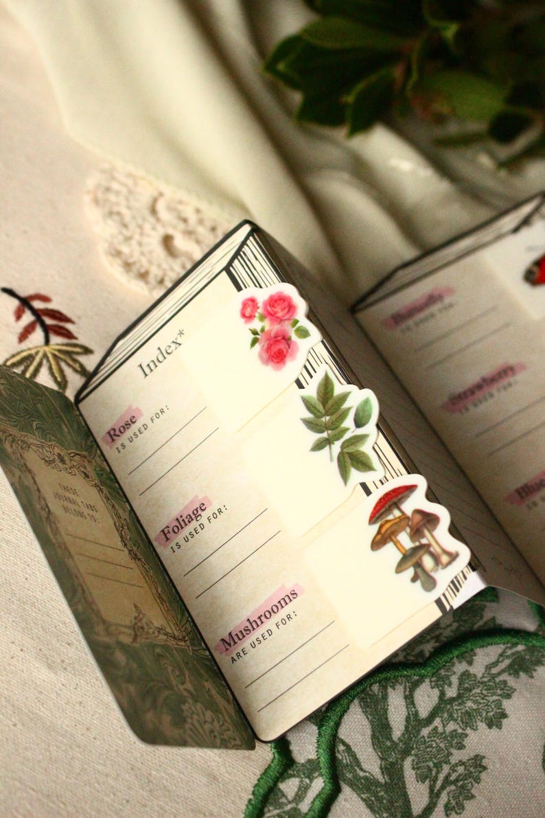 Pode incluir: Di&aacute;rio aberto com adesivos decorativos de flores e cogumelos. A p&aacute;gina do &iacute;ndice apresenta as palavras "Rose", "Foliage" e "Mushrooms" com linhas para anota&ccedil;&otilde;es. O di&aacute;rio est&aacute; aberto em uma superf&iacute;cie creme.