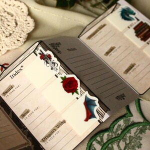 Pode incluir: Caderno aberto com cart&otilde;es de &iacute;ndice com ilustra&ccedil;&otilde;es de uma espada, rosa e asa de drag&atilde;o. As p&aacute;ginas do caderno t&ecirc;m linhas para anota&ccedil;&otilde;es. Adesivos decorativos e um desenho de &aacute;rvore bordada em verde e branco tamb&eacute;m s&atilde;o vis&iacute;veis.