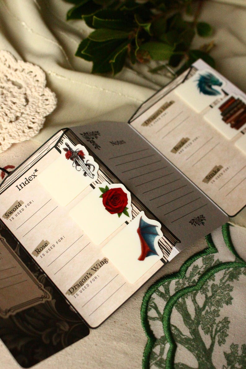 Pode incluir: Caderno aberto com cart&otilde;es de &iacute;ndice com ilustra&ccedil;&otilde;es de uma espada, rosa e asa de drag&atilde;o. As p&aacute;ginas do caderno t&ecirc;m linhas para anota&ccedil;&otilde;es. Adesivos decorativos e um desenho de &aacute;rvore bordada em verde e branco tamb&eacute;m s&atilde;o vis&iacute;veis.