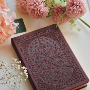 Puede incluir: Un diario de color burdeos con un diseño floral en relieve en la portada. El diario rectangular presenta un patrón detallado. Flores rosas y paniculata blanca están dispuestas alrededor del diario sobre una superficie blanca.