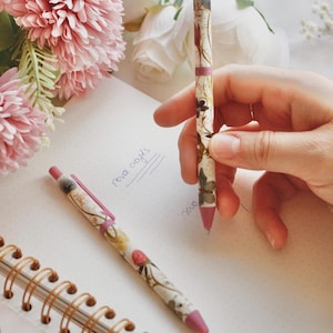 May include: Two floral-patterned pens with pink accents are on a dotted notebook. One pen is held by a hand. The pens feature a design of flowers and leaves. Pink and white flowers are in the background.