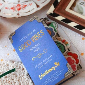 Pode incluir: Um di&aacute;rio azul com letras douradas "Little Book of Good Vibes" e cantos decorativos. Cont&eacute;m 60 marcadores de p&aacute;gina autocolantes. Autocolantes florais e smiley coloridos tamb&eacute;m s&atilde;o vis&iacute;veis.