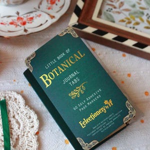 Pode incluir: Um "Little Book of Botanical Journal Tabs" verde-azulado com letras douradas e detalhes decorativos nos cantos. O livro cont&eacute;m 60 marcadores de p&aacute;gina autocolantes. Uma ch&aacute;vena de ch&aacute; e arte emoldurada no fundo.