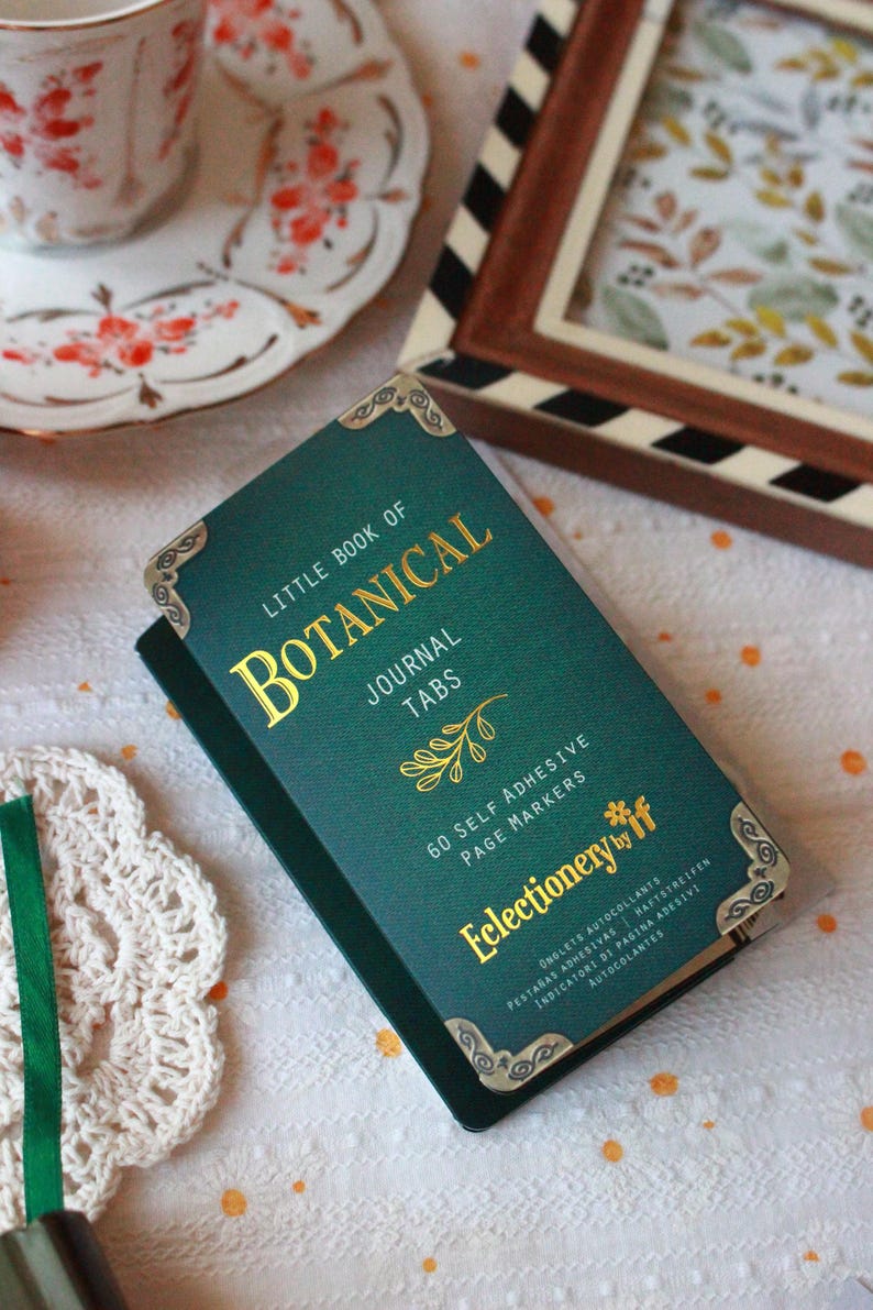 Pode incluir: Um "Little Book of Botanical Journal Tabs" verde-azulado com letras douradas e detalhes decorativos nos cantos. O livro cont&eacute;m 60 marcadores de p&aacute;gina autocolantes. Uma ch&aacute;vena de ch&aacute; e arte emoldurada no fundo.
