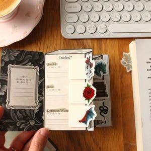Pode incluir: Um di&aacute;rio aberto com abas decorativas com uma espada, uma rosa e uma asa de drag&atilde;o. O di&aacute;rio inclui o texto "Estas abas do di&aacute;rio pertencem a:" e est&aacute; sobre uma superf&iacute;cie de madeira. Uma ch&aacute;vena de caf&eacute; e um teclado est&atilde;o no fundo.