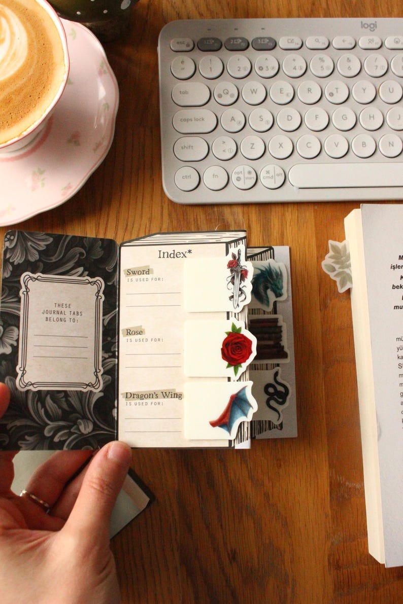 Pode incluir: Um di&aacute;rio aberto com abas decorativas com uma espada, uma rosa e uma asa de drag&atilde;o. O di&aacute;rio inclui o texto "Estas abas do di&aacute;rio pertencem a:" e est&aacute; sobre uma superf&iacute;cie de madeira. Uma ch&aacute;vena de caf&eacute; e um teclado est&atilde;o no fundo.