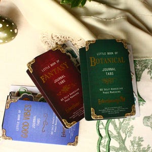 Pode incluir: Um conjunto de abas de di&aacute;rio com bordas douradas decorativas. As abas s&atilde;o rotuladas como "Little Book of Fantasy", "Little Book of Botanical" e "Little Book of Good Vibes". Cada conjunto cont&eacute;m 60 marcadores de p&aacute;gina autoadesivos.