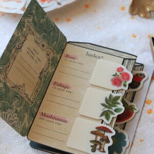 Pode incluir: Di&aacute;rio aberto com adesivos florais e de cogumelos. A capa apresenta um design floral verde e o texto "THESE JOURNAL TABS BELONG TO:". O &iacute;ndice lista "Rose", "Foliage" e "Mushrooms".