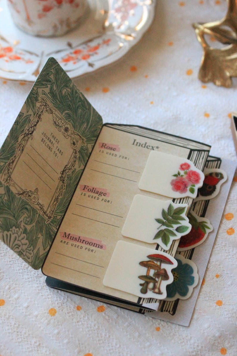 Pode incluir: Di&aacute;rio aberto com adesivos florais e de cogumelos. A capa apresenta um design floral verde e o texto "THESE JOURNAL TABS BELONG TO:". O &iacute;ndice lista "Rose", "Foliage" e "Mushrooms".