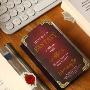 May include: A burgundy "Little Book of Fantasy" journal tab set with gold accents and a rose bookmark rests on a wooden surface near a keyboard and a book. The journal tabs are self-adhesive.