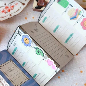 Pode incluir: Di&aacute;rio aberto com abas decorativas com ilustra&ccedil;&otilde;es de uma flor, uma seta e um cora&ccedil;&atilde;o. As abas s&atilde;o rotuladas com frases como "Coffee" e "Orange Flower". O di&aacute;rio tem p&aacute;ginas pautadas.