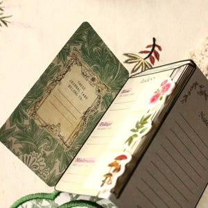 Pode incluir: Di&aacute;rio aberto com abas de &iacute;ndice decorativas com temas florais e de cogumelos. O di&aacute;rio tem uma capa verde com um design floral e o texto "These Journal Tabs Belong To:". As p&aacute;ginas s&atilde;o pautadas.