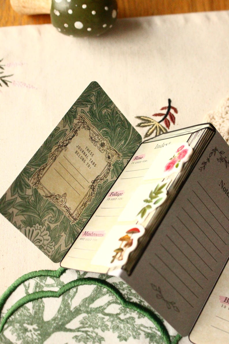 Pode incluir: Di&aacute;rio aberto com abas de &iacute;ndice decorativas com temas florais e de cogumelos. O di&aacute;rio tem uma capa verde com um design floral e o texto "These Journal Tabs Belong To:". As p&aacute;ginas s&atilde;o pautadas.