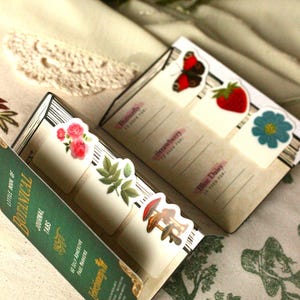 Pode incluir: Um conjunto de abas de di&aacute;rio bot&acirc;nico com ilustra&ccedil;&otilde;es florais e de cogumelos. As abas est&atilde;o presas a um pequeno livro verde com o texto "LITTLE BOOK OF BOTANICAL JOURNAL TABS". As abas apresentam imagens de uma borboleta, morango e margarida azul.