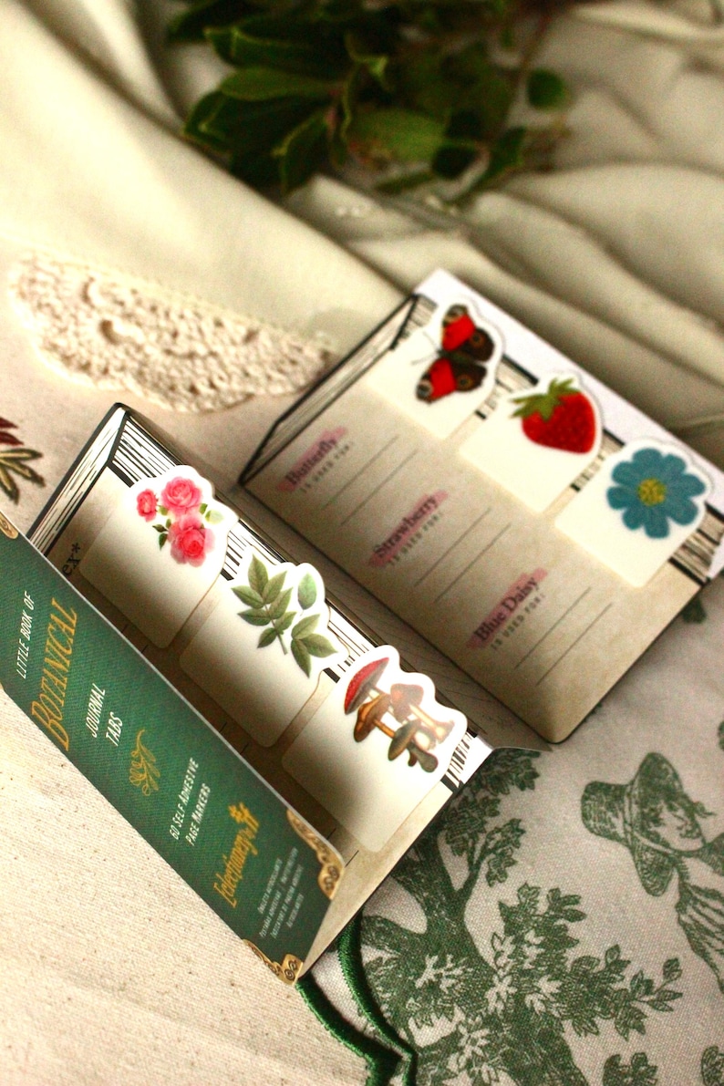 Pode incluir: Um conjunto de abas de di&aacute;rio bot&acirc;nico com ilustra&ccedil;&otilde;es florais e de cogumelos. As abas est&atilde;o presas a um pequeno livro verde com o texto "LITTLE BOOK OF BOTANICAL JOURNAL TABS". As abas apresentam imagens de uma borboleta, morango e margarida azul.