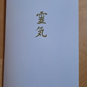 Könnte beinhalten: Ein weißes Papier mit den japanischen Schriftzeichen für Reiki in Gold gedruckt. Das Papier ist rechteckig und scheint ein Buchumschlag oder ein Flyer zu sein. Der Hintergrund ist eine hellbraune Holzoberfläche.