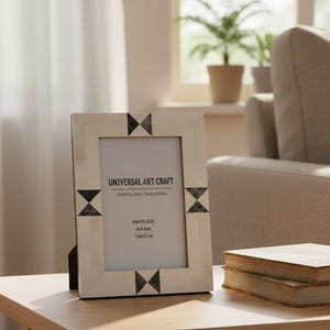 Può includere: Cornice fotografica rettangolare in legno chiaro con dettagli geometrici neri. La cornice mostra il testo «UNIVERSAL ART CRAFT» ed è progettata per una foto di 10x15 cm. È su un tavolo di legno, vicino a due libri.