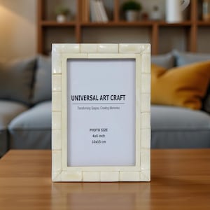 Puede incluir: Un marco de fotos rectangular con un diseño de mosaico de color crema. El marco contiene un papel blanco con el texto "UNIVERSAL ART CRAFT" y "PHOTO SIZE 4x6 inch, 10x15 cm". El marco está sobre una mesa de madera.