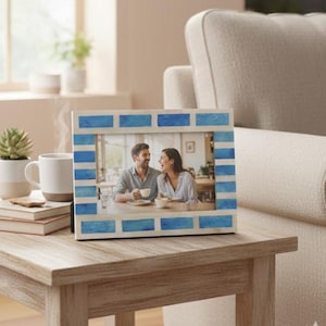 May include: A rectangular photo frame with a blue and white brick pattern border. The frame holds a photograph of a couple. The frame is on a light wood table, with a coffee cup and a succulent plant in the background.