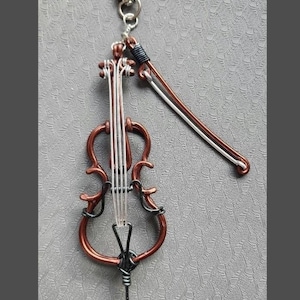 May include: A handcrafted wire art keychain featuring a miniature cello with a bow. The cello is made of copper-colored wire with silver strings and black accents. The bow is silver and copper-colored. A silver clasp is attached.