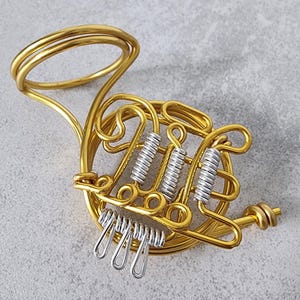 Puede incluir: Una escultura de trompa francesa en miniatura hecha de alambre dorado. El instrumento presenta resortes de válvula y llaves de dedo plateados, con un diseño detallado. La escultura es un artículo decorativo, probablemente para los amantes de la música.