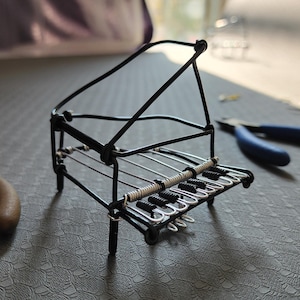 May include: A miniature black wire piano with a detailed keyboard and strings. The small sculpture is set on a textured gray surface, with a pair of blue pliers in the background. The piano's design includes a music stand.