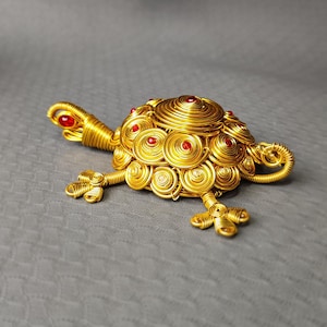 May include: A handcrafted golden wire turtle sculpture with red accents. The turtle's shell is made of concentric circles, and it has red gems on its back and head. The legs and head are detailed with wire. The sculpture is on a gray surface.