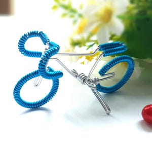 Puede incluir: Una pequeña escultura de bicicleta hecha de metal plateado y alambre azul brillante. La bicicleta presenta un marco, ruedas y manillar detallados, con el alambre azul agregando un toque de color. La escultura se encuentra sobre un fondo suave y borroso.