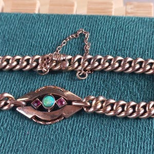 May include: A gold-coloured bracelet with a chain link design. The bracelet features a central decorative element with a turquoise stone and two red stones. The bracelet is displayed on a teal surface.