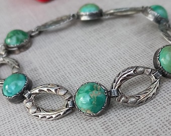 Op de afbeelding: Een vintage zilveren armband met ovale en ronde turkooiskleurige stenen. De armband heeft een sierlijk filigraanontwerp op de schakels, wat een klassieke en elegante uitstraling creëert. De stenen hebben een levendige groene kleur.
