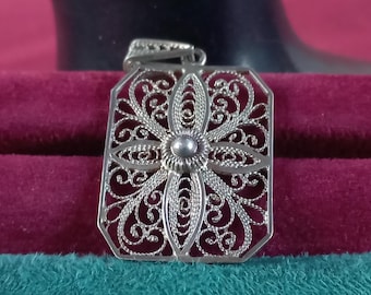 Könnte beinhalten: Silberner Filigrananhänger mit offenem Design. Der Anhänger hat ein zentrales Blumenelement, das von komplizierten Wirbelmustern umgeben ist. Der rechteckige Rahmen hat eine dekorative Öse für eine Kette. Der Anhänger wird vor einem bordeauxroten und türkisfarbenen Hintergrund präsentiert.
