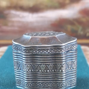 Puede incluir: Una pequeña caja octogonal de plata con un acabado desgastado. La tapa y los lados presentan intrincados patrones florales y geométricos en relieve, incluyendo motivos de estrellas repetitivos y diseños de hojas estilizadas alrededor del cuerpo.