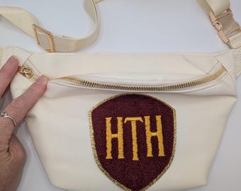 May include: Cream-coloured fanny pack with a gold zip and strap. The pack features a burgundy shield-shaped patch with the gold letters "HTH". The strap has a gold buckle.