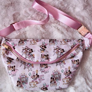 Puede incluir: Riñonera rosa con un estampado repetido de Mickey y Minnie Mouse en varias atracciones de parques temáticos. La bolsa tiene una cremallera rosa, una correa ajustable rosa y una hebilla dorada.