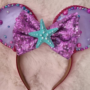 May include: A headband featuring two mouse ears, a sequined bow, and a turquoise starfish. The ears are purple with a beaded trim and iridescent accents. The bow is made of purple sequins. The headband is red.